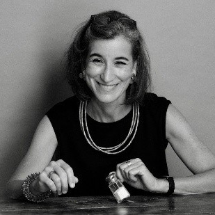A person sits at a table holding a small jar, wearing a sleeveless top, layered necklaces, a bracelet, and a watch.