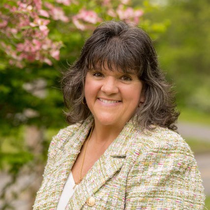 Person in a textured jacket smiles outdoors near pink flowering trees.