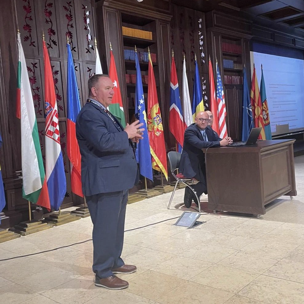 A speaker addresses an audience beside seated panelists with national flags displayed across the stage.
