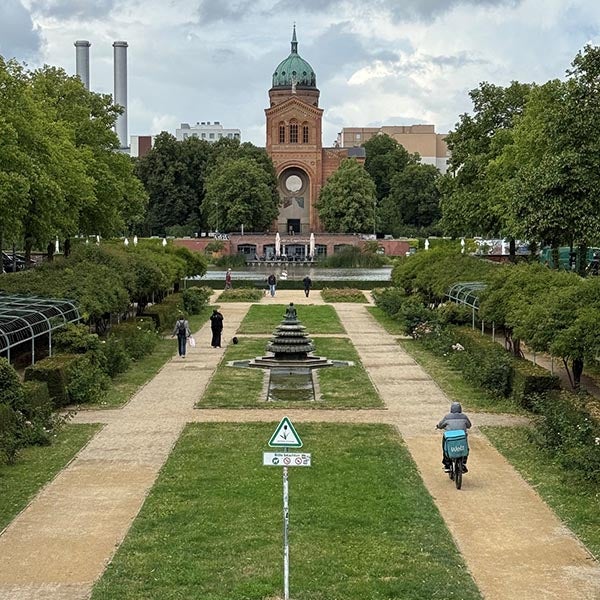 Formal garden with a central fountain, walking paths, trees, cyclists, and a brick tower in the distance.