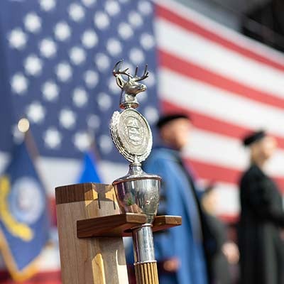 Norwich University ceremonial mace stands before a large U.S. flag and people in academic regalia.
