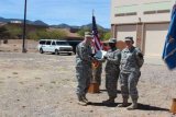 Three service members in camouflage uniforms stand beside U.S. and military flags during an outdoor presentation ceremony.