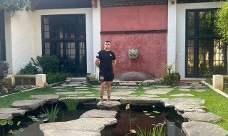 Sam Nucup standing on stepping stones over a pond in front of a building with black-framed windows and a red wall.