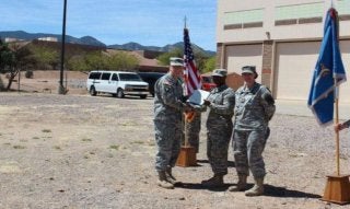 Three service members in camouflage uniforms stand beside U.S. and military flags during an outdoor presentation ceremony.