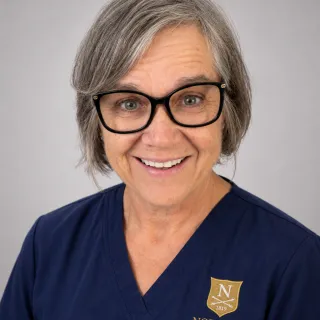 Person with short gray hair and glasses wears navy Norwich University School of Nursing scrubs.