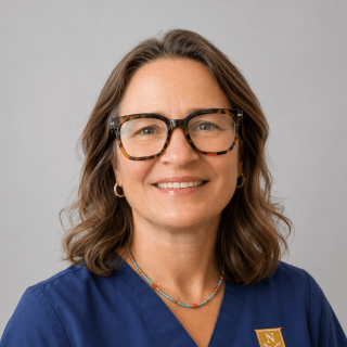 A person wearing glasses and blue Norwich University scrubs poses against a gray background.