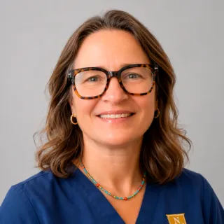 A person wearing glasses and blue Norwich University scrubs poses against a gray background.