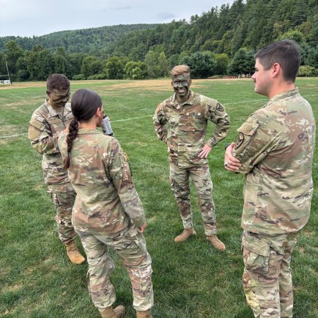 Four people in camouflage uniforms stand in a circle talking on a grassy field with trees in the background.