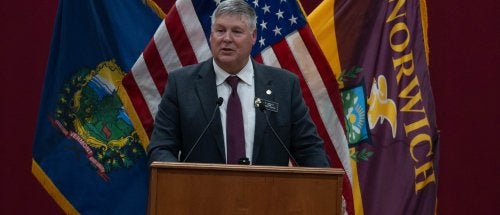 Person speaks at a Norwich University podium with Vermont, U.S., and Norwich flags behind.