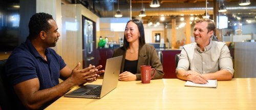 Three people sit at a table with a laptop and notebook during a meeting in a shared workspace.