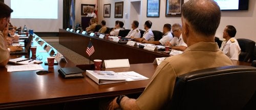 Military officers attend a conference room presentation with slides.