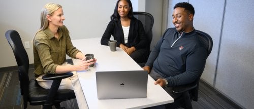 Three people sit around a table with a laptop and mugs during a meeting.
