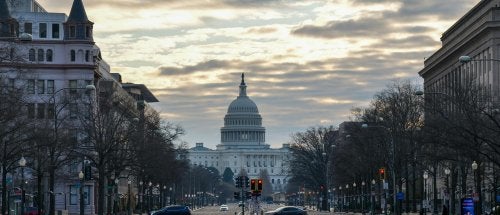 U.S. Capitol stands at the end of a broad avenue lined with buildings and bare trees under a cloudy sky.