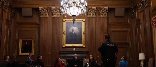 Police officer stands before seated people in a wood-paneled courtroom with a chandelier and framed portraits.