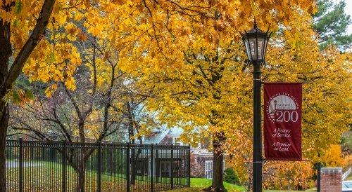 A black metal fence runs along a walkway past a lamp post with a red banner reading "Norwich University 200" in front of vivid yellow autumn trees.