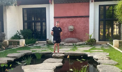 Sam Nucup standing on stepping stones over a pond in front of a building with black-framed windows and a red wall.
