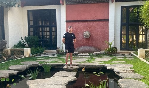 Sam Nucup standing on stepping stones over a pond in front of a building with black-framed windows and a red wall.