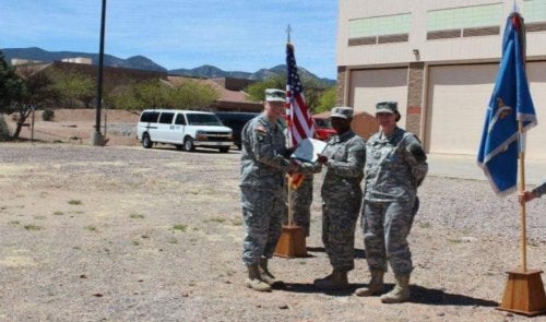 Three service members in camouflage uniforms stand beside U.S. and military flags during an outdoor presentation ceremony.