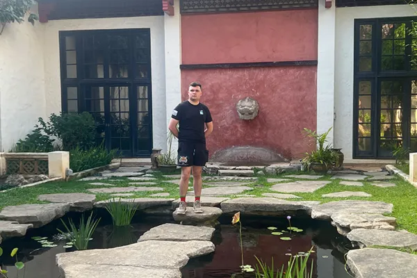Sam Nucup standing on stepping stones over a pond in front of a building with black-framed windows and a red wall.