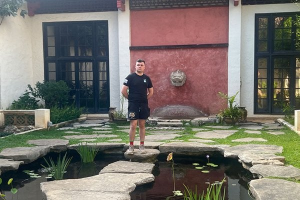 Sam Nucup standing on stepping stones over a pond in front of a building with black-framed windows and a red wall.
