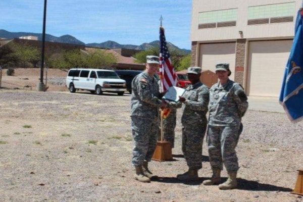 Three service members in camouflage uniforms stand beside U.S. and military flags during an outdoor presentation ceremony.